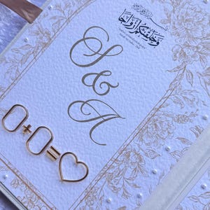 Op de afbeelding: Een witte huwelijksuitnodiging met gouden bloemen- en parelaccenten. Het ontwerp bevat de letters "S & A" in elegant schrift, samen met een gouden metalen vergelijking: 0+0=♡. De uitnodiging bevat ook Arabisch schrift.