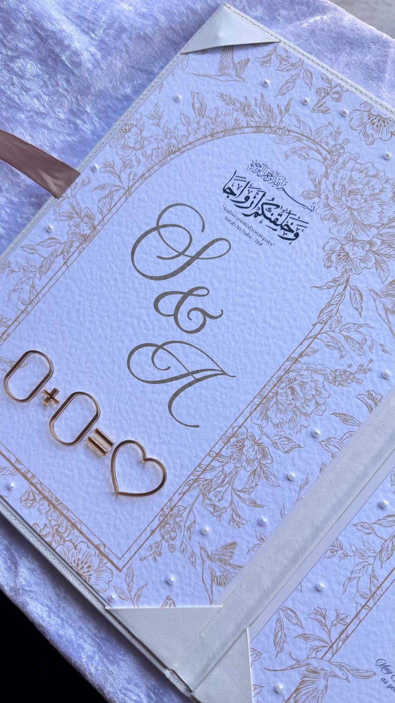 Op de afbeelding: Een witte huwelijksuitnodiging met gouden bloemen- en parelaccenten. Het ontwerp bevat de letters "S & A" in elegant schrift, samen met een gouden metalen vergelijking: 0+0=♡. De uitnodiging bevat ook Arabisch schrift.