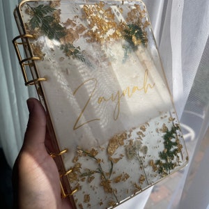 May include: A white resin-bound journal with gold leaf and pressed flowers. The journal has a gold ring binding and the name "Zaynah" is written in gold script on the front cover.