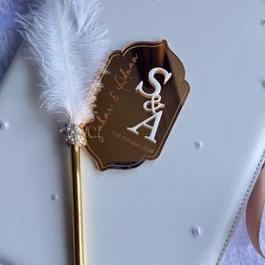 Op de afbeelding: Een wit trouwgastenboek met gouden accenten en een decoratieve pen. De pen heeft een witte veer en parelversieringen. Een gouden spiegelachtige tag toont de namen "Sahar & Adnan" en de datum "12 februari 2026".