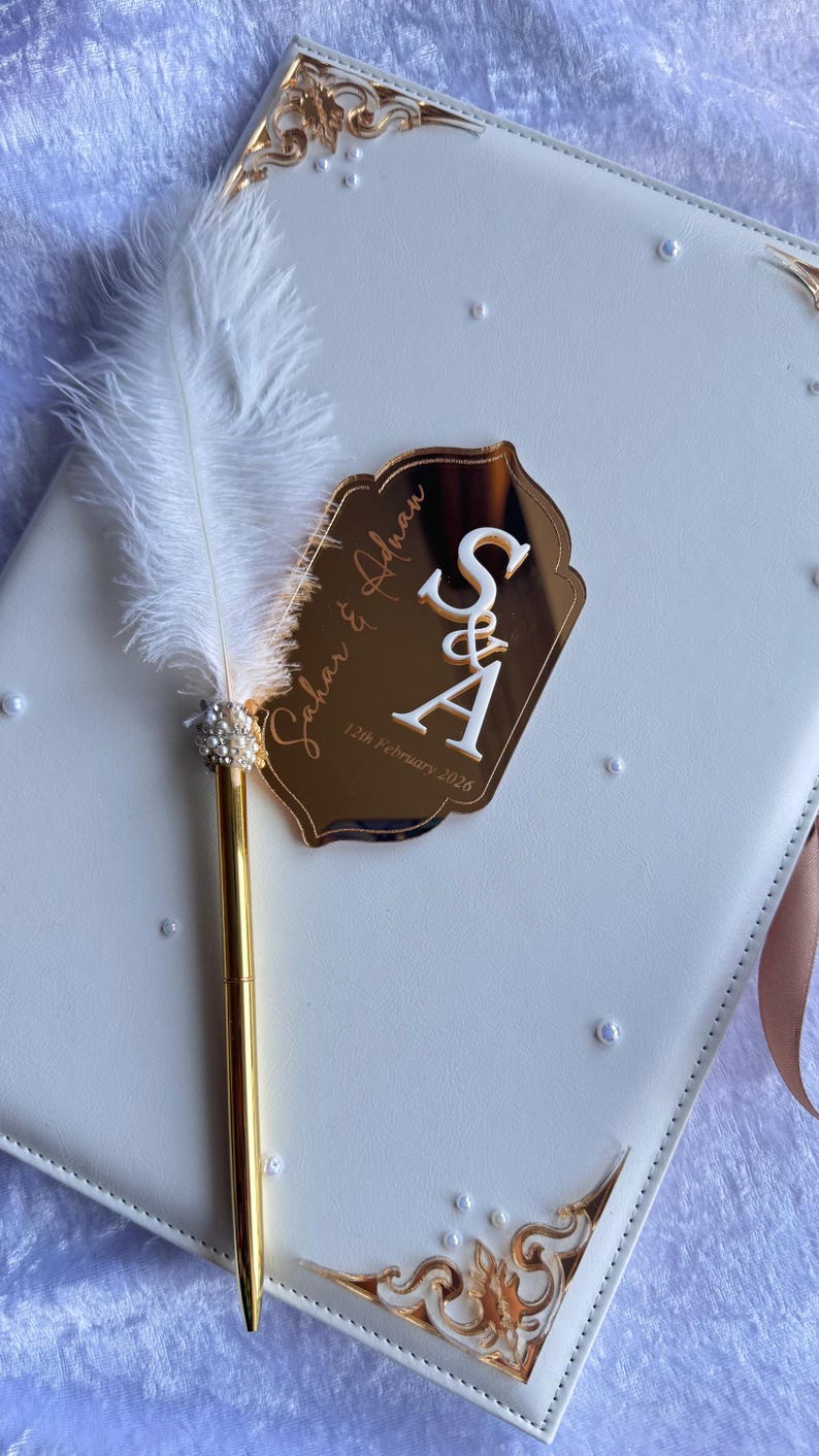 Op de afbeelding: Een wit trouwgastenboek met gouden accenten en een decoratieve pen. De pen heeft een witte veer en parelversieringen. Een gouden spiegelachtige tag toont de namen "Sahar & Adnan" en de datum "12 februari 2026".