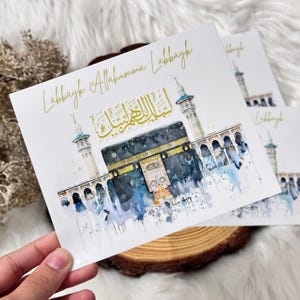 Puede incluir: Una tarjeta blanca con una pintura de acuarela de la Kaaba en La Meca, Arabia Saudita. La tarjeta tiene letras doradas que dicen "Labbayk Allahumma Labbayk".