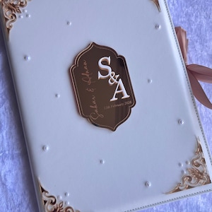 Op de afbeelding: Een wit trouwgastenboek met gouden details en een decoratief embleem. Het embleem toont de initialen "S&A" en de datum "12th February 2026". De hoeken zijn versierd met gouden filigraan.