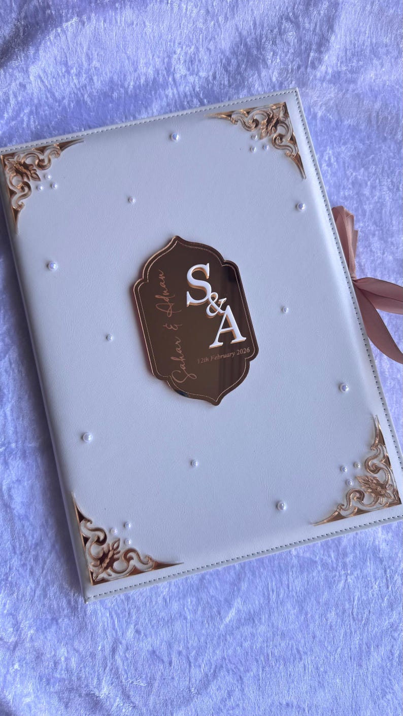 Op de afbeelding: Een wit trouwgastenboek met gouden details en een decoratief embleem. Het embleem toont de initialen "S&A" en de datum "12th February 2026". De hoeken zijn versierd met gouden filigraan.