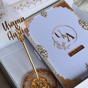 May include: A white box with a gold-trimmed book inside. The book has a white cover with gold lettering that says "Usama & Afsana" and "U&A" with a gold date tag that says "14.02.2025". A gold pen and stand are in the box.