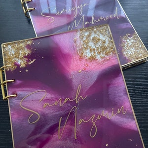May include: Two purple and white resin-bound journals with gold leaf accents and gold lettering. The top journal has the name "Sumiya Mahmood" written on it. The bottom journal has the name "Sanah Nazmin" written on it.