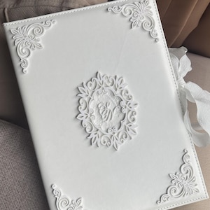 May include: A white rectangular album with a smooth cover featuring ornate, raised floral corner accents and a central oval medallion containing intricate lace-like detailing and embossed script lettering. A white ribbon is attached to the side.