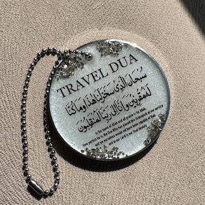 Op de afbeelding: Een ronde, transparante sleutelhanger met de zwarte tekst "TRAVEL DUA". De sleutelhanger heeft Arabische tekst en Engelse tekst. Zilveren glitter accentueert de randen en er is een zilveren kogelketting bevestigd.