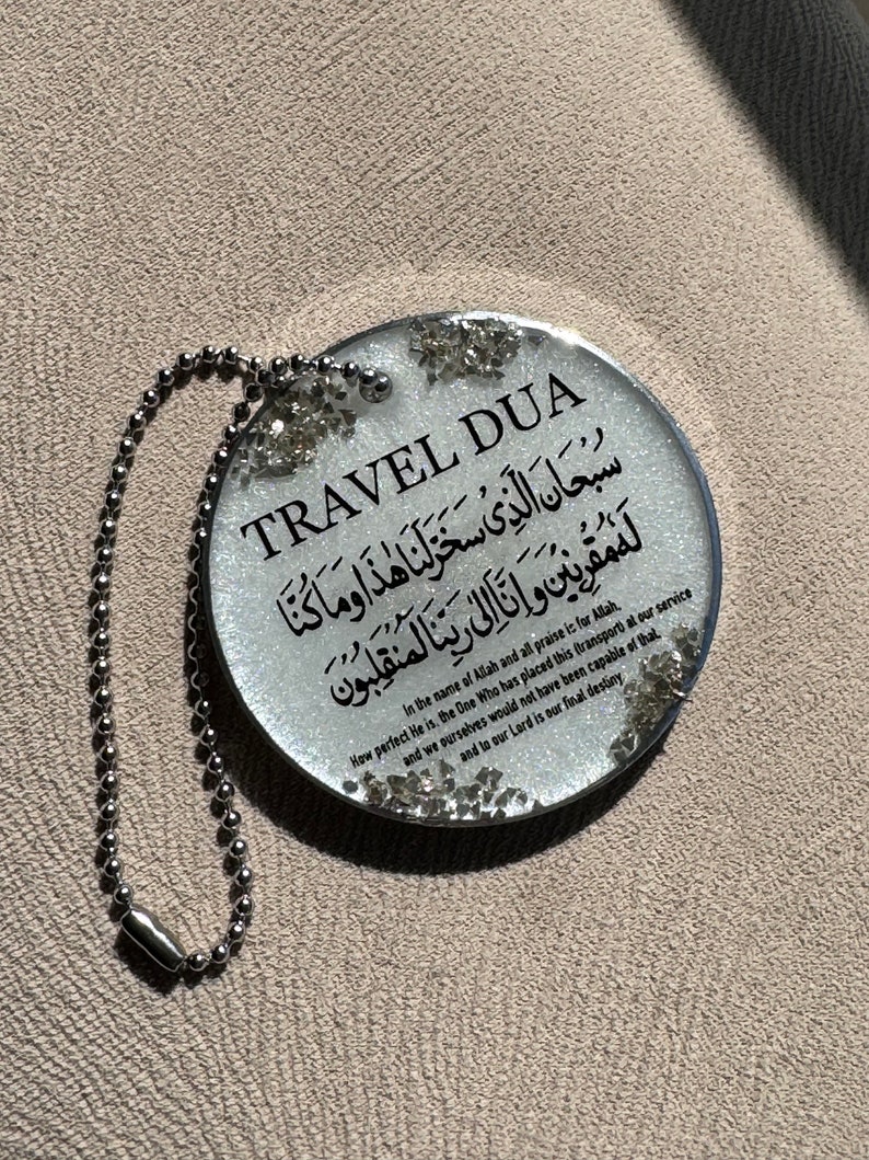 Op de afbeelding: Een ronde, transparante sleutelhanger met de zwarte tekst "TRAVEL DUA". De sleutelhanger heeft Arabische tekst en Engelse tekst. Zilveren glitter accentueert de randen en er is een zilveren kogelketting bevestigd.