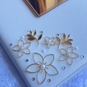 以下が含まれることがあります： 金色の花の装飾と金色の鏡のようなタグが付いた白いノート。タグには「Aqeb & Maliha」の名前が刻まれています。ノートは、金のワイヤーフラワー、葉、真珠、ラインストーンで飾られています。