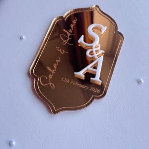 Op de afbeelding: Een goudkleurig acryl huwelijksbedankje met de namen "Sahar & Adnan" en de datum "12 februari 2026" gegraveerd. De initialen "S&A" zijn wit, met een decoratieve ampersand. Het bedankje ligt op een witte ondergrond.