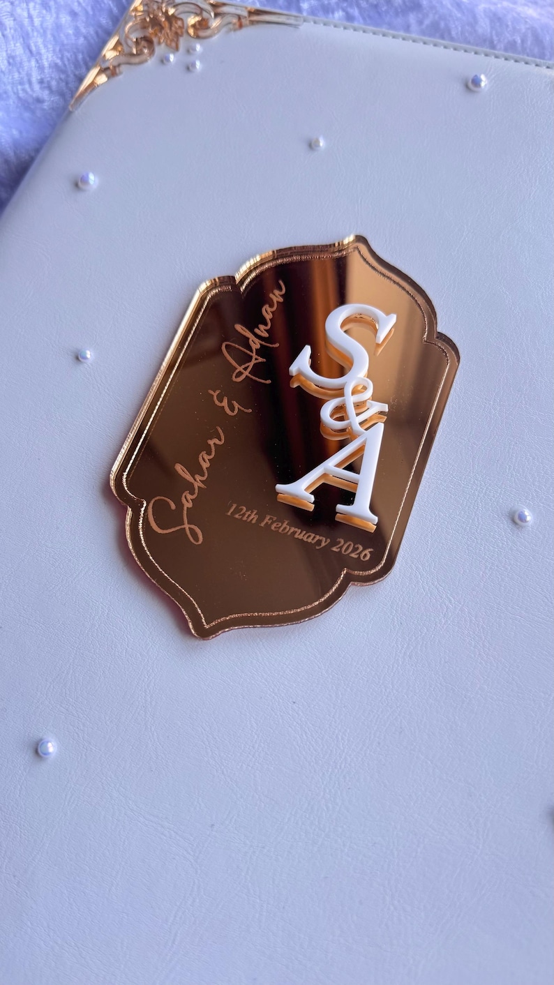 Op de afbeelding: Een goudkleurig acryl huwelijksbedankje met de namen "Sahar & Adnan" en de datum "12 februari 2026" gegraveerd. De initialen "S&A" zijn wit, met een decoratieve ampersand. Het bedankje ligt op een witte ondergrond.
