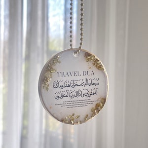 Op de afbeelding: Een ronde, heldere hars auto-hanger met bladgoud accenten en een gouden ketting. De hanger heeft Arabisch schrift en de Engelse tekst "TRAVEL DUA". De achtergrond is een zacht, wazig wit.