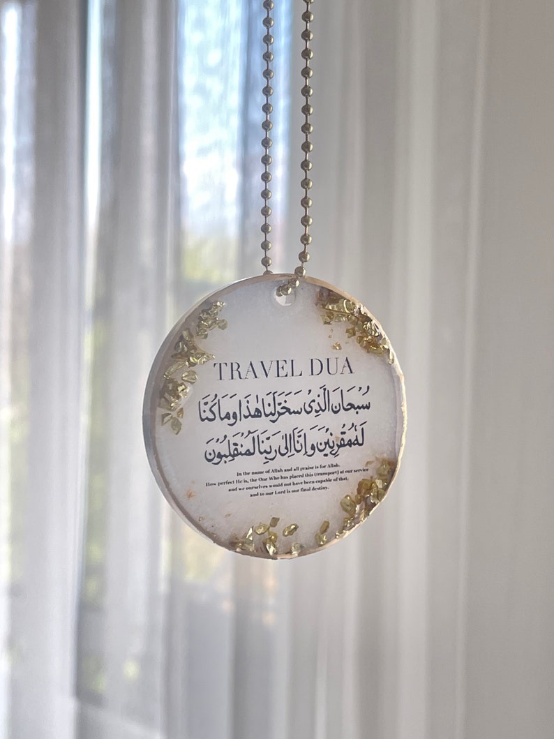Op de afbeelding: Een ronde, heldere hars auto-hanger met bladgoud accenten en een gouden ketting. De hanger heeft Arabisch schrift en de Engelse tekst "TRAVEL DUA". De achtergrond is een zacht, wazig wit.
