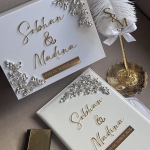 May include: A white rectangular box and a matching white book, both featuring gold script text reading Sobhan & Madina, the date 4TH OCTOBER 2026, and clear crystal floral embellishments. A white feather pen with a gold stand and S&M topper.