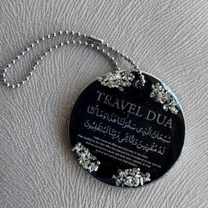 Op de afbeelding: Een zwarte, ronde reistag met zilveren accenten en een zilveren kogelketting. De tag bevat de woorden "TRAVEL DUA" en Arabisch schrift, samen met Engelse tekst. Het is een religieus item.