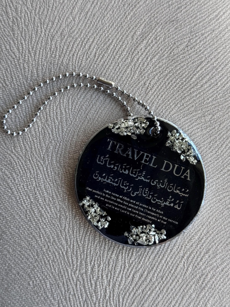 Op de afbeelding: Een zwarte, ronde reistag met zilveren accenten en een zilveren kogelketting. De tag bevat de woorden "TRAVEL DUA" en Arabisch schrift, samen met Engelse tekst. Het is een religieus item.