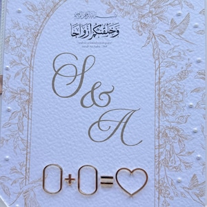 Op de afbeelding: Een trouwuitnodiging met gouden bloemen- en scriptdetails. De uitnodiging bevat de tekst "S & A" en de vergelijking "0 + 0 = ♡". De bovenkant van de uitnodiging heeft Arabisch schrift en de Engelse tekst "And we created you in pairs".