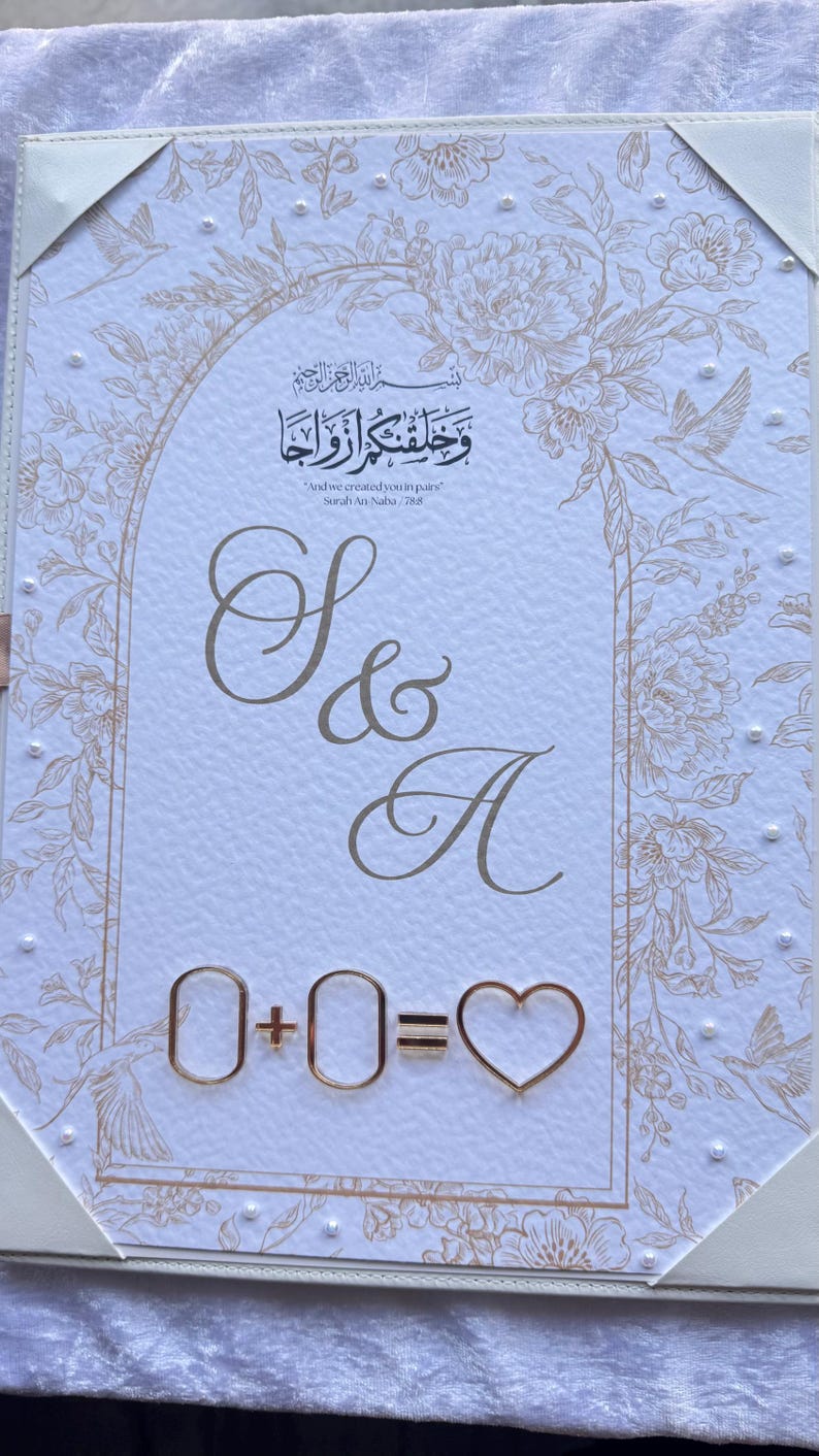 Op de afbeelding: Een trouwuitnodiging met gouden bloemen- en scriptdetails. De uitnodiging bevat de tekst "S & A" en de vergelijking "0 + 0 = ♡". De bovenkant van de uitnodiging heeft Arabisch schrift en de Engelse tekst "And we created you in pairs".