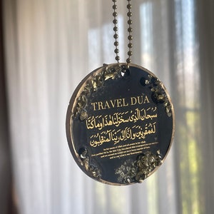 Op de afbeelding: Zwarte ronde hanger met gouden tekst en accenten, hangend aan een bronzen kogelketting. De hanger toont de woorden "TRAVEL DUA" en Arabisch schrift. De randen zijn versierd met goudkleurige fragmenten.