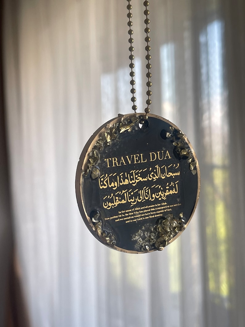 Op de afbeelding: Zwarte ronde hanger met gouden tekst en accenten, hangend aan een bronzen kogelketting. De hanger toont de woorden "TRAVEL DUA" en Arabisch schrift. De randen zijn versierd met goudkleurige fragmenten.