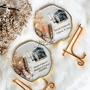 May include: Two white resin coasters with gold trim featuring a printed image of the Kaaba in Mecca, Saudi Arabia. Each coaster has dried flowers and a personalised message. The message on the left coaster reads "Umrah Mubarak Shamim & Nadeem 2021/1443". The message on the right coaster reads "Umrah Mubarak Safwan & Sara".
