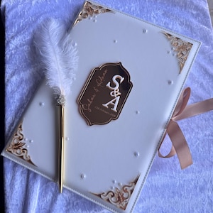 Op de afbeelding: Een wit gastenboek met gouden details en een gouden pen met een veer. Het boek heeft een gouden plaatje met de initialen "S & A" en de datum "12 februari 2028". Een lichtbruin lint is om het boek gebonden.