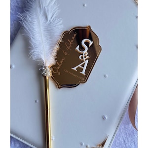 Op de afbeelding: Een wit trouwgastenboek met gouden details, een gouden pen met een witte veer en een gouden spiegelplaat met de initialen "S&A" en de datum "12 februari 2026". Het boek heeft parelversieringen en decoratieve hoeken.