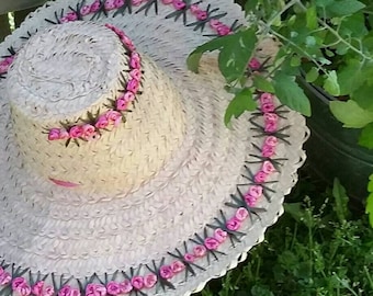 May include: A large, wide-brimmed straw hat with a pink flower trim. The hat is woven with a light brown straw and has a black ribbon around the crown.
