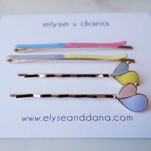 Op de afbeelding: Set van zes gouden haarclips met kleurrijke emaille accenten. Twee clips hebben een tweekleurig ontwerp met blauw en roze emaille. De andere vier clips hebben hartvormige accenten met geel, roze en blauw emaille.