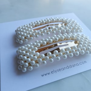 Könnte beinhalten: Zwei goldene Haarklammern, die mit weißen Perlen besetzt sind. Die Klammern sind rechteckig und haben einen dezenten Glanz. Der Text "www.elyseanddana.com" ist auf einer weißen Oberfläche unter den Klammern aufgedruckt.