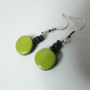 Pode incluir: Um par de brincos feitos à mão com um pendente circular verde brilhante. O pendente está ligado a uma série de pequenas contas cinza escuro e a um gancho de orelha prateado. Design simples e elegante.