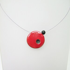 Può includere: Una collana minimalista con un ciondolo rosso vivo con un piccolo foro e una perla nera. Il ciondolo è sospeso a un filo sottile, creando un effetto fluttuante. Il gioiello è esposto su sfondo bianco. Il diametro del ciondolo è di circa 2,5 cm.