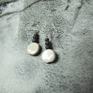 May include: A pair of handmade earrings featuring a combination of black and off-white elements. Each earring has a silver hook, a cluster of small black beads, and a large, round, off-white ceramic bead. The earrings are displayed against a textured gray background.