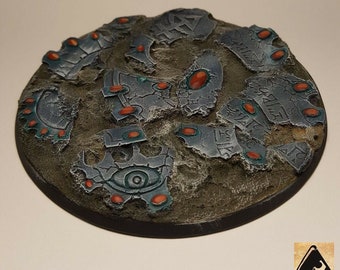 May include: A round, gray, and brown miniature base with a textured surface. The base is decorated with silver and blue metallic paint, and features several red gems. The base is designed to be used for tabletop games.