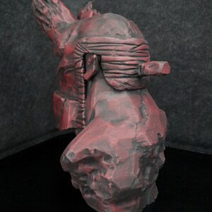 Puede incluir: Un busto esculpido en rojo y negro de una persona con una venda en los ojos. El busto est&aacute; hecho de un material rugoso y texturizado y tiene un dise&ntilde;o detallado.