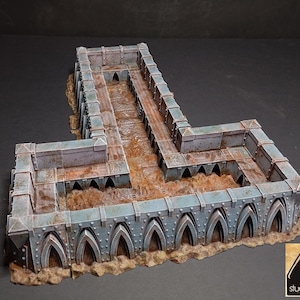 May include: A detailed, L-shaped miniature terrain piece with a weathered, industrial aesthetic. The structure features a blue-grey metal finish with rust-colored accents, arched openings, and a textured base, perfect for tabletop gaming.