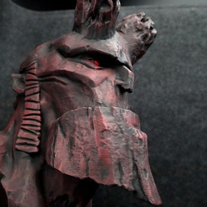 Puede incluir: Un busto de escultura de piedra roja y negra de una figura con cuernos y un ojo rojo brillante. La escultura est&aacute; detallada con l&iacute;neas texturizadas y una superficie irregular de tipo roca.