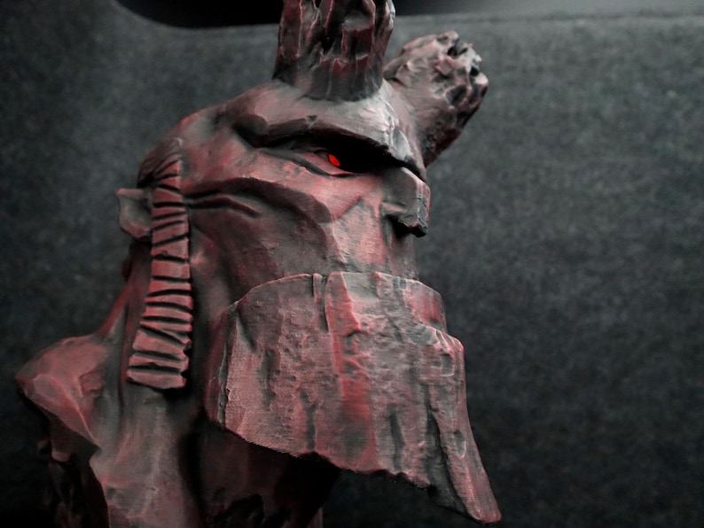 Puede incluir: Un busto de escultura de piedra roja y negra de una figura con cuernos y un ojo rojo brillante. La escultura est&aacute; detallada con l&iacute;neas texturizadas y una superficie irregular de tipo roca.