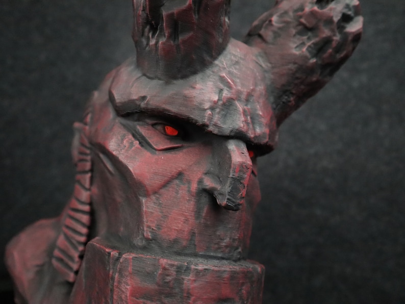 Puede incluir: Primer plano de una escultura de piedra gris oscuro y roja de una criatura con una gran corona dentada y ojos rojos brillantes.