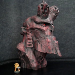 Puede incluir: Un busto rojo y rocoso detallado de un personaje con un solo ojo rojo. El busto est&aacute; hecho de un material texturizado y tiene una corona rocosa y dentada. El busto est&aacute; sobre una superficie negra.