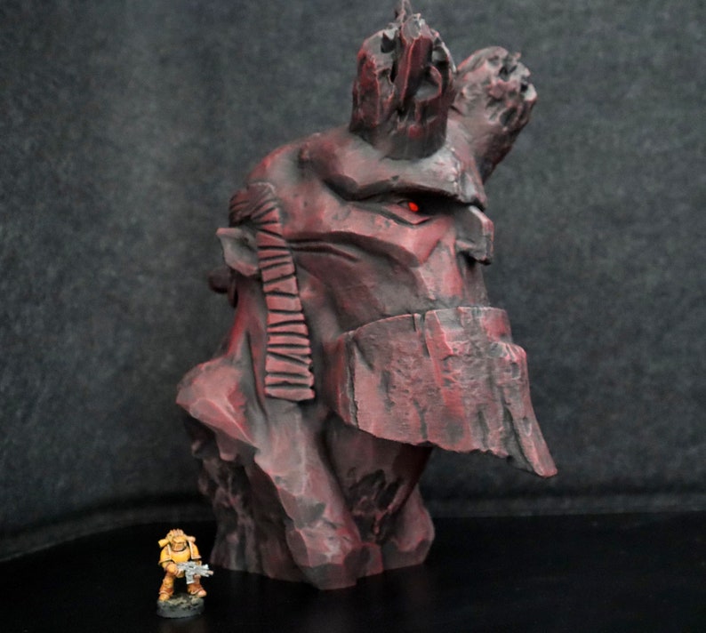 Puede incluir: Un busto rojo y rocoso detallado de un personaje con un solo ojo rojo. El busto est&aacute; hecho de un material texturizado y tiene una corona rocosa y dentada. El busto est&aacute; sobre una superficie negra.