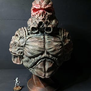 Puede incluir: Busto detallado de una figura monstruosa con una apariencia texturizada y desgastada. La escultura presenta una cabeza grotesca con ojos rojos brillantes y un torso musculoso. El busto está montado sobre una base circular.