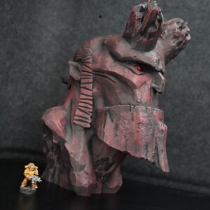 Puede incluir: Una escultura de busto detallada en rojo y negro que representa a un personaje con una gran corona dentada. La escultura est&aacute; hecha de un material texturizado que se asemeja a la roca. Una peque&ntilde;a figura pintada de amarillo de un soldado se encuentra frente a la escultura.