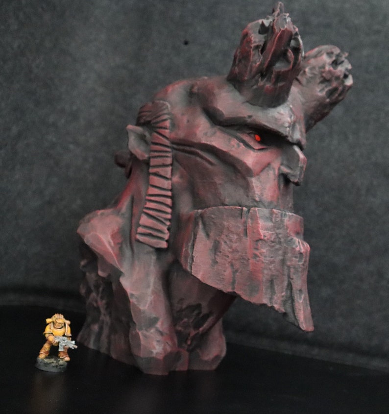 Puede incluir: Una escultura de busto detallada en rojo y negro que representa a un personaje con una gran corona dentada. La escultura est&aacute; hecha de un material texturizado que se asemeja a la roca. Una peque&ntilde;a figura pintada de amarillo de un soldado se encuentra frente a la escultura.