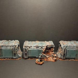 Puede incluir: Tres contenedores de almacenamiento en miniatura de color verde azulado desgastado con daños de batalla, incluyendo agujeros de bala y secciones desmoronadas. Una pequeña figura se encuentra frente al contenedor central. Diseñado para juegos de mesa.