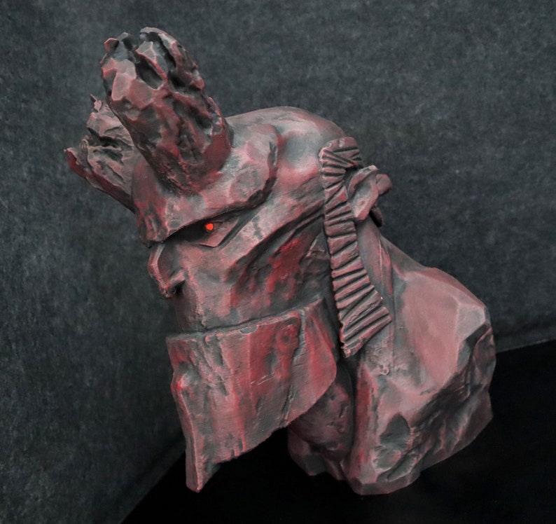 Puede incluir: Una escultura de piedra roja y gris de un demonio con cuernos y un solo ojo rojo. La escultura es detallada y texturizada, asemej&aacute;ndose a una formaci&oacute;n rocosa.