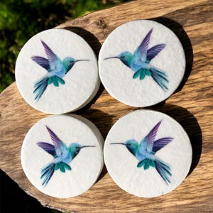 Könnte beinhalten: Vier runde Untersetzer mit einem Kolibri-Design. Jeder Untersetzer hat einen weißen Hintergrund mit einer detaillierten Illustration eines Kolibris in Blau-, Lila- und Grüntönen. Die Untersetzer werden auf einer Holzoberfläche präsentiert.