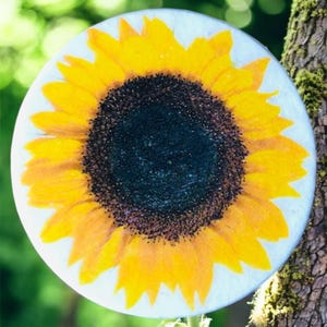 Może przedstawiać: Okrągły przedmiot dekoracyjny z żywym wzorem słonecznika. Słonecznik ma jasne żółte płatki otaczające ciemnobrązowy środek. Przedmiot jest umieszczony na rozmytym zielonym tle, sugerującym otoczenie zewnętrzne.