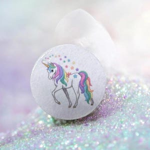 Può includere: Un bottone rotondo bianco con un'illustrazione di unicorno colorato. L'unicorno ha un corno dorato, una criniera e una coda arcobaleno e accenti di stelle. Lo sfondo ha una consistenza morbida e scintillante. Il bottone misura circa 3 cm di diametro.